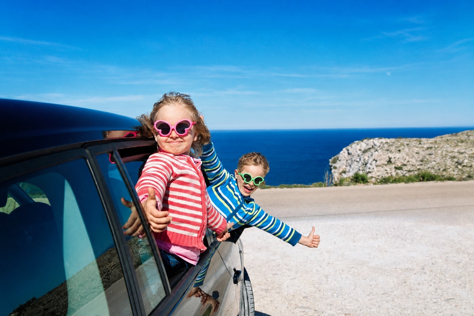 Two happy children wearing colourful sunglasses and sweaters leaning out of a car window during a coastal road trip, with the ocean in the background.